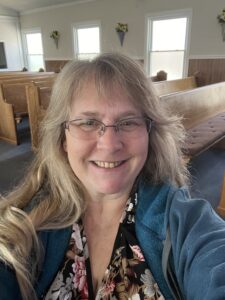 A picture of Tiffani smiling at the camera. She is wearing wire-rimmed purple glasses, a flowered navy dress, and a medium blue jacket. She is standing in her church with the pews visible in the background.